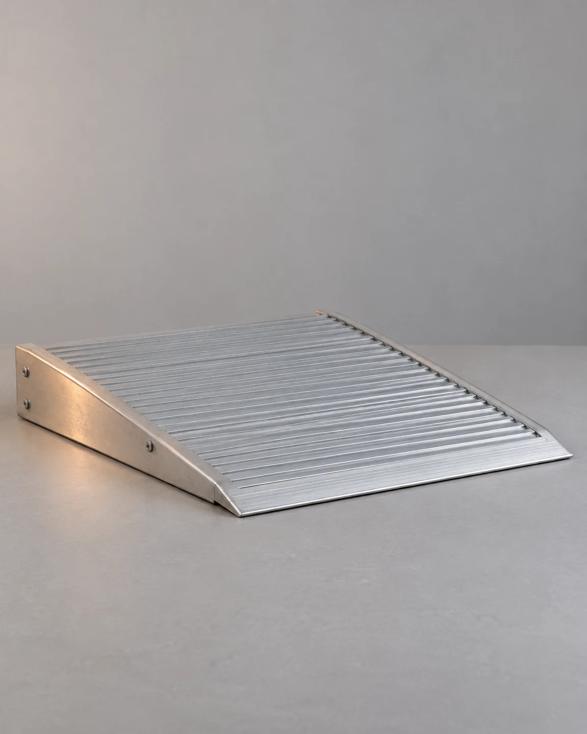 A silver ribbed aluminum threshold ramp rests diagonally on a matte gray tabletop, with its wedge-shaped side and thin front lip visible against an uncluttered…