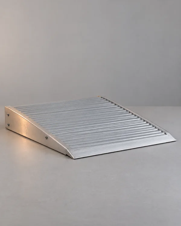A silver ribbed aluminum threshold ramp rests diagonally on a matte gray tabletop, with its wedge-shaped side and thin front lip visible against an uncluttered…