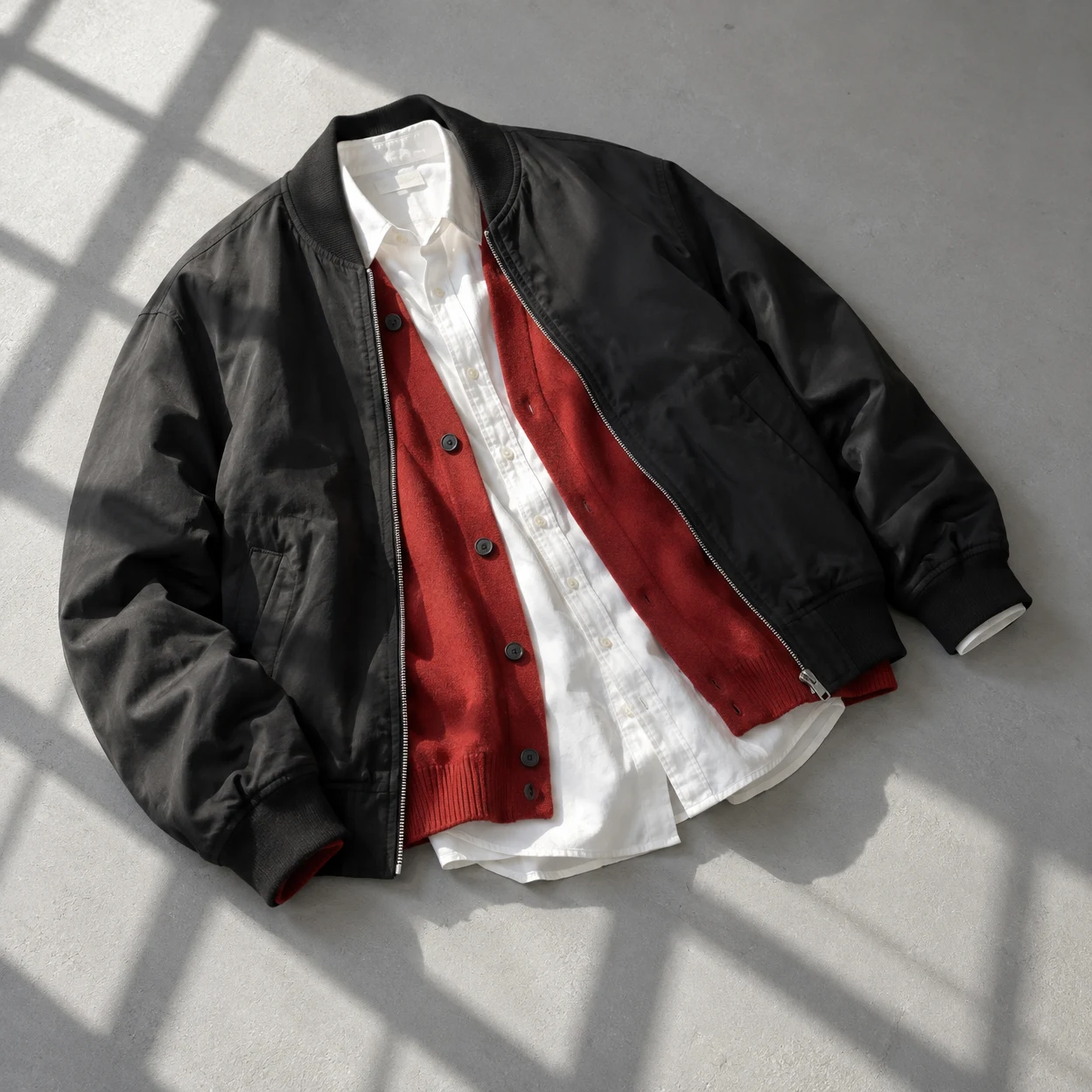 Overhead studio photograph of a black bomber jacket layered over a vermilion buttoned cardigan and white shirt, arranged diagonally on a pale grey floor with…