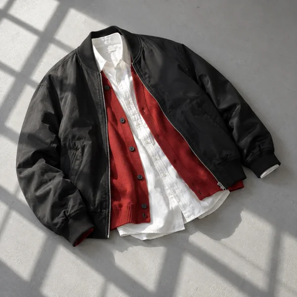 Overhead studio photograph of a black bomber jacket layered over a vermilion buttoned cardigan and white shirt, arranged diagonally on a pale grey floor with…
