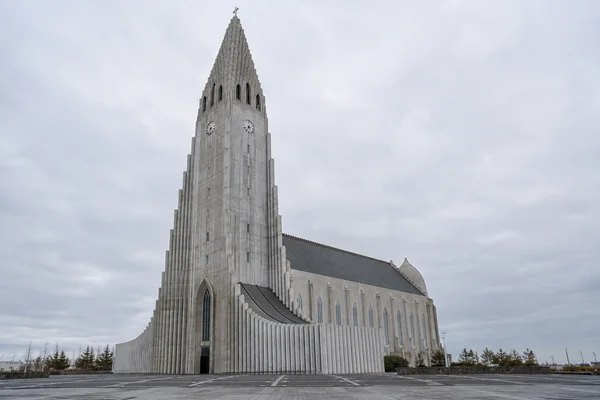 A pale concrete church with a tall stepped tower and cross stands beside a broad paved forecourt beneath a cloudy grey sky.