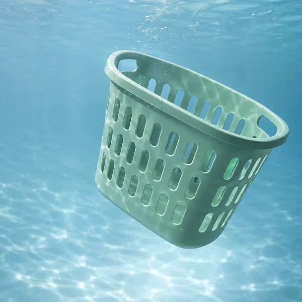 A mint-green plastic laundry basket with rounded ventilation slots and integrated handles floats diagonally in blue water, with small bubbles around its rim…