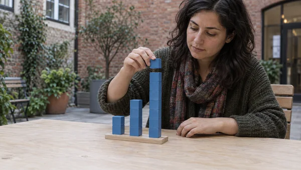 A woman sits at a pale wooden table on a brick courtyard terrace, holding a blue cube above the tallest of three blue-cube columns arranged on a rectangular…