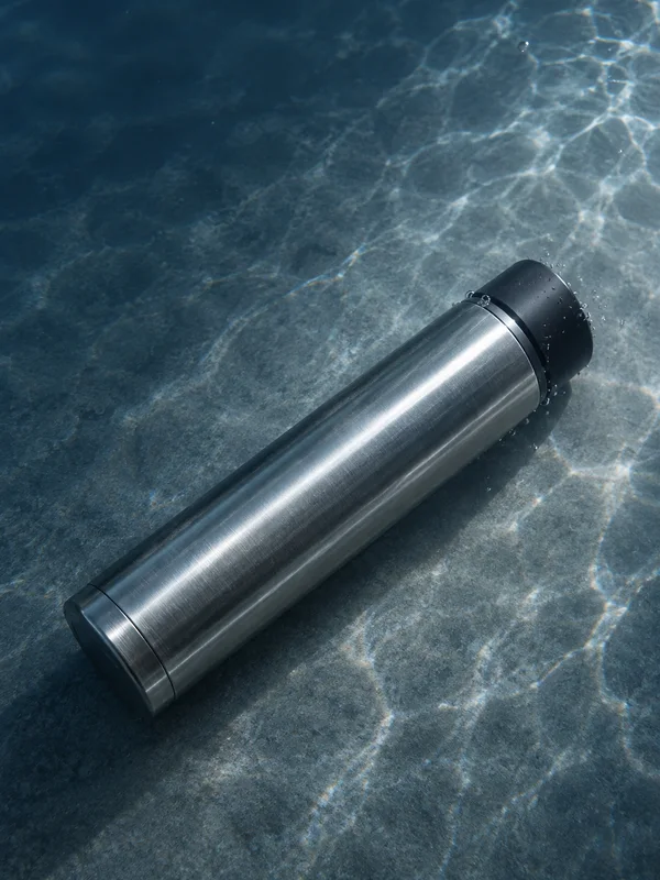 A brushed stainless-steel thermos with a matte-black cap lies diagonally on a textured slate-grey platform underwater, with bright caustic patterns and bubbles…
