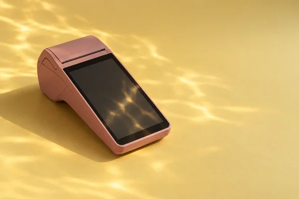 A muted rose point-of-sale terminal rests diagonally on a butter-yellow surface, with golden rippled reflections across its dark screen and the surrounding…