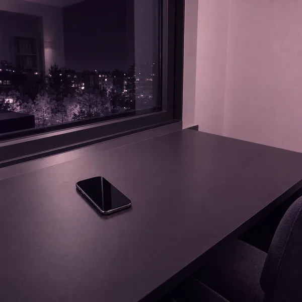 A black-screen smartphone lies diagonally on a broad dark desk beside a large night window, with a pale rose wall and city lights visible beyond the glass.