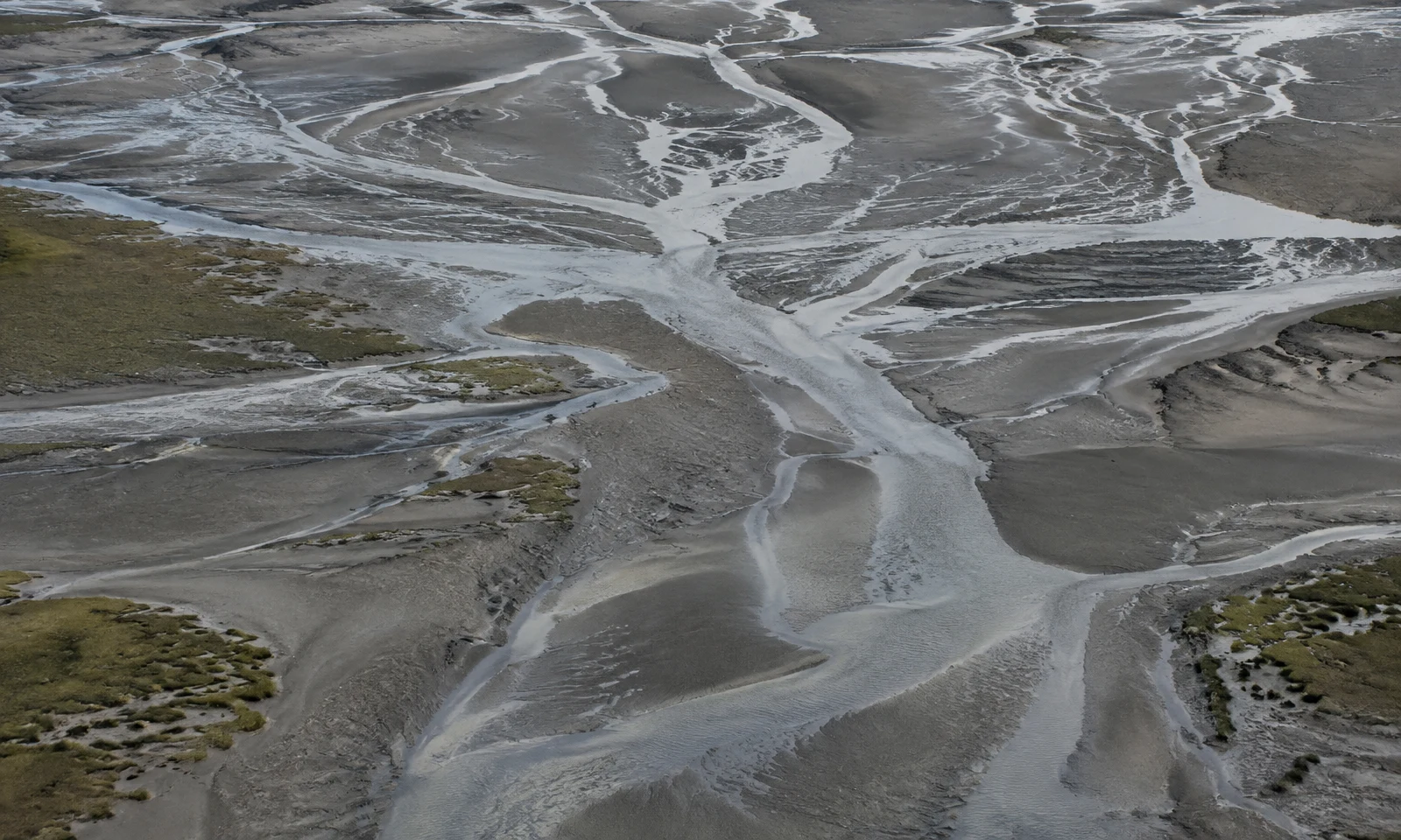 A high-angle view across an estuary at low tide, where silvery braided channels cross grey-brown mudflats with sparse olive saltmarsh patches.