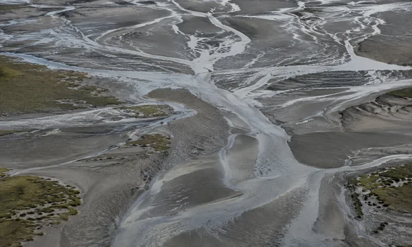 A high-angle view across an estuary at low tide, where silvery braided channels cross grey-brown mudflats with sparse olive saltmarsh patches.