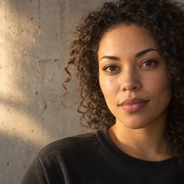 A person with dark curly hair in a charcoal crewneck looks toward the camera in a tight portrait, with warm light on one side of the face and a textured…