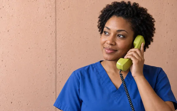 A nurse in cobalt-blue scrubs holds a chartreuse telephone handset to her ear, looking toward the open left side with a small smile.
