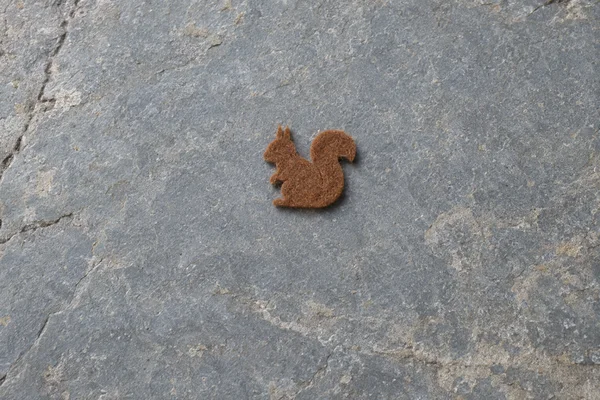 A small cinnamon-brown felt cutout shaped like a seated squirrel rests on a blue-grey stone surface, viewed from above.