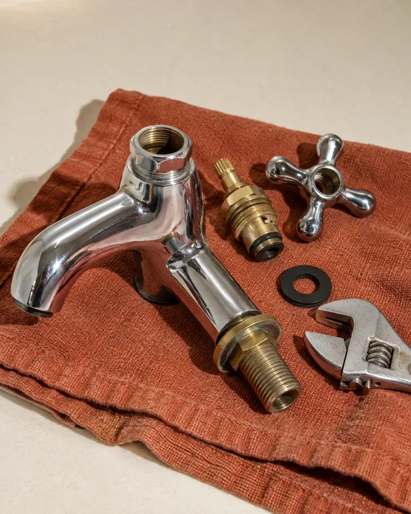 A detached chrome pillar tap lies diagonally on a rust-coloured cotton towel on a cream worktop, with its brass valve assembly, chrome crosshead handle, black…