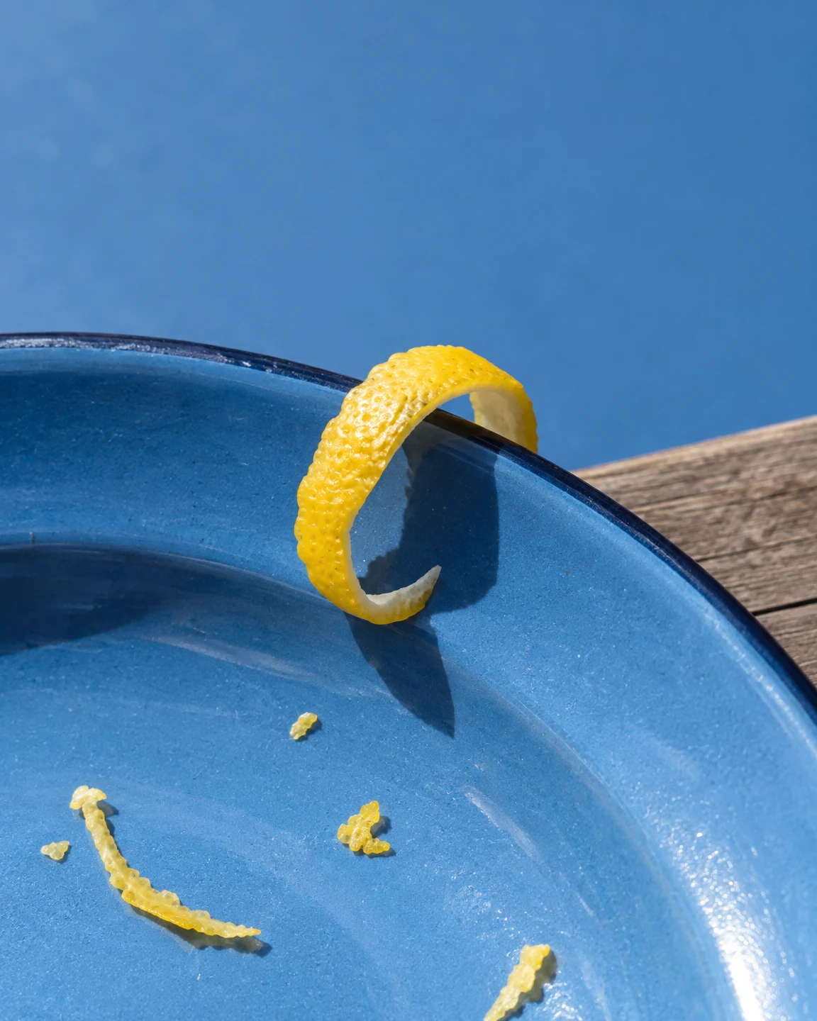 A thick yellow lemon-peel curl loops over the rim of a blue enamel plate, with several loose peel pieces scattered across it; a weathered wooden surface shows…