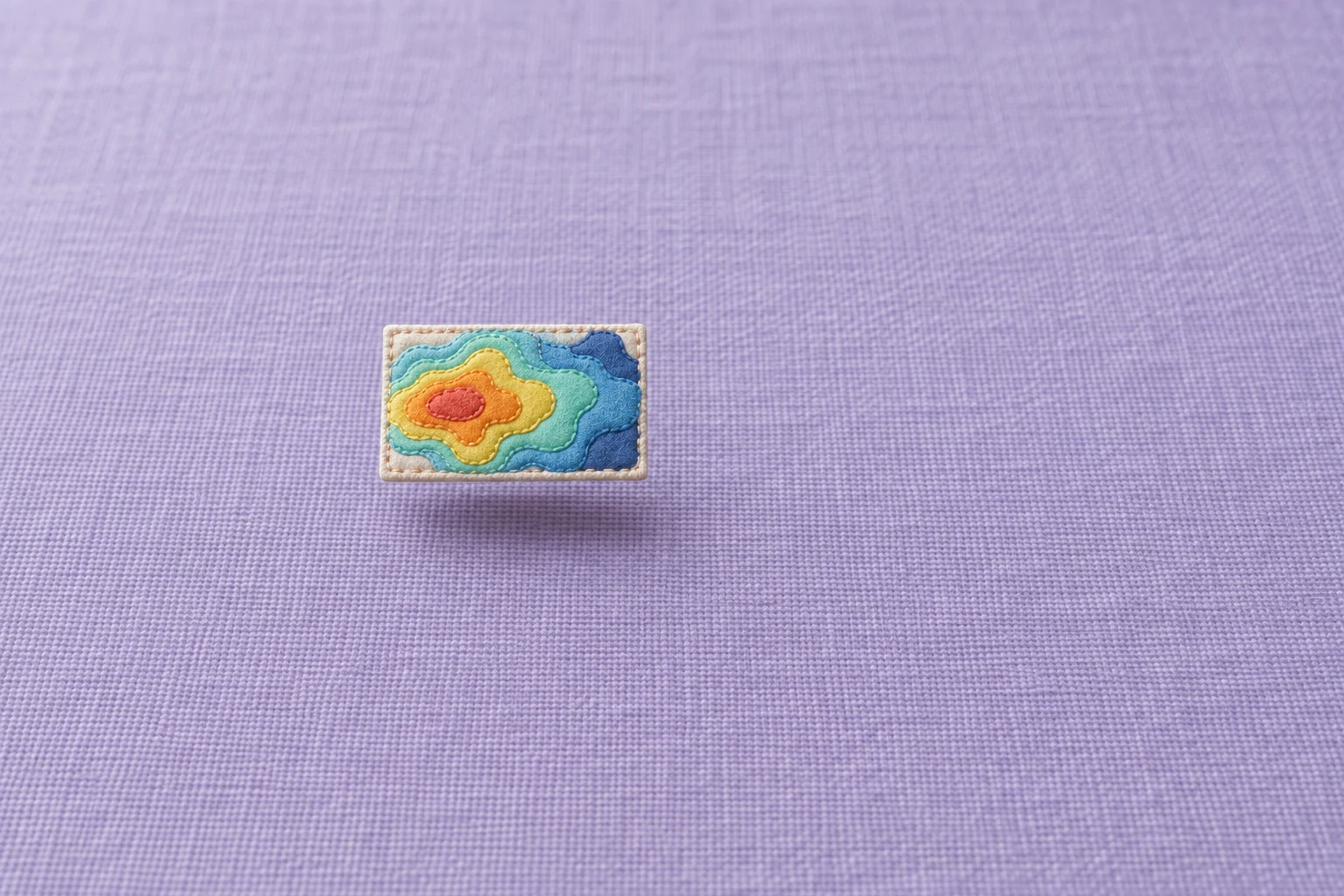 A small rectangular quilt tile with concentric coral, orange, yellow, aqua and blue fabric zones floats above a lavender woven surface, casting a faint shadow…