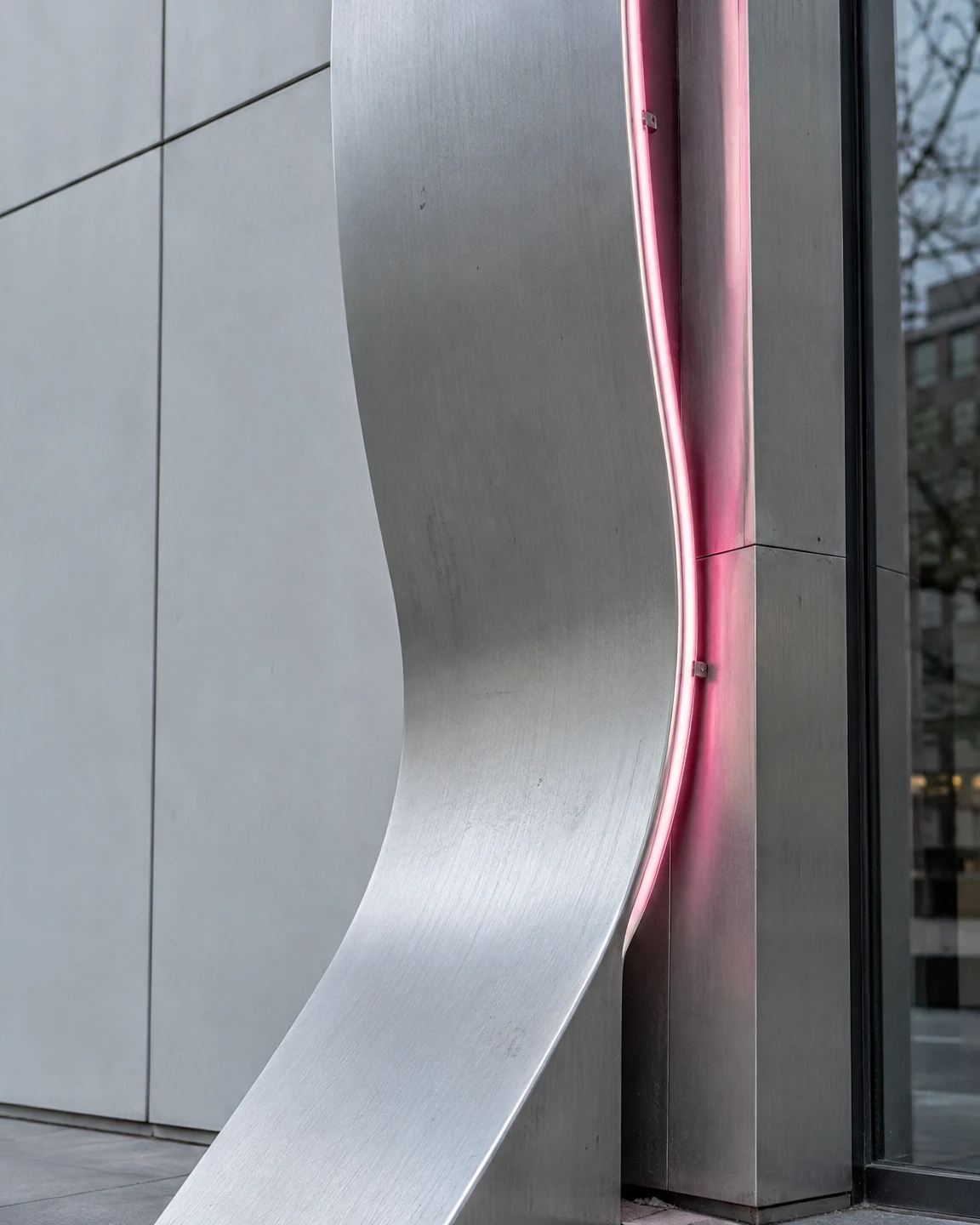 A broad curved brushed-aluminum facade fin fills the center beside a narrow pink neon edge, with pale gray panels to the left and a dark glazed frontage at…