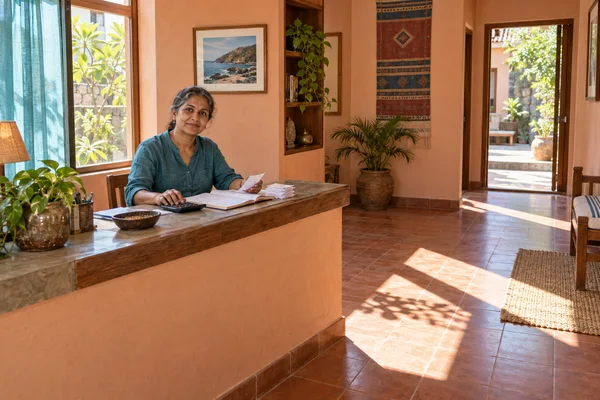 A woman in a teal shirt sits behind a wooden reception counter, handling papers beside an open ledger and calculator in a sunlit peach-walled room with…
