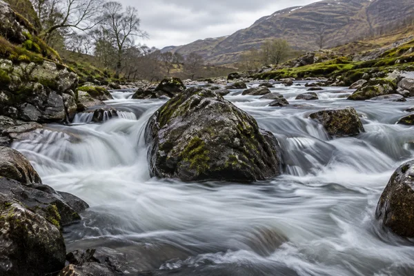 A boulder-strewn upland river rushes around a large moss-flecked dark boulder in the foreground, with silky white water, wet stones, wooded banks, and muted…