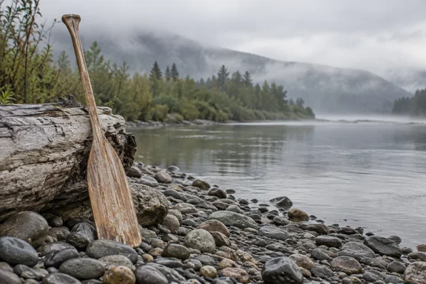A weathered wooden canoe paddle leans against a large bleached driftwood log on a pebbly riverbank, with a broad misty river and wooded banks behind it.