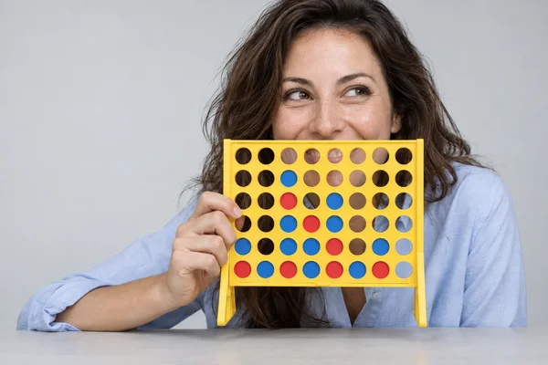 A woman in a pale-blue shirt laughs while holding a yellow perforated game grid in front of her mouth at a light-grey studio table, looking off-camera.