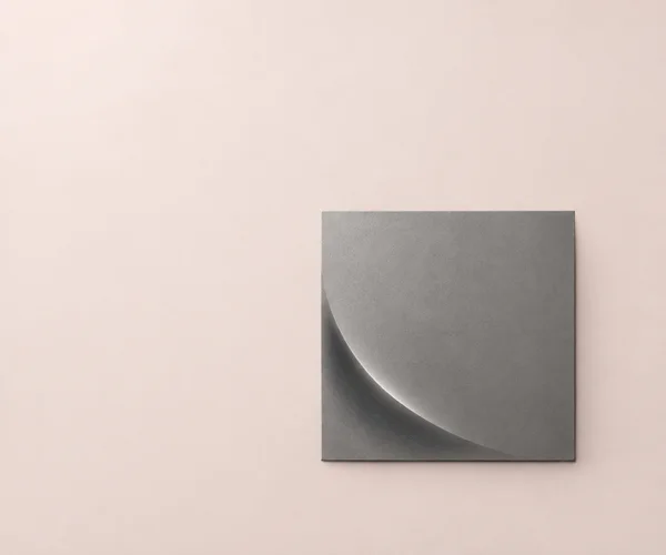 A textured pewter-grey square tile sits below centre and slightly right on a pale blush background, with a broad curved shadow and highlight sweeping across…