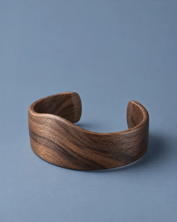 A broad open walnut cuff bracelet with a gently wavy rim rests on a matte blue-grey surface, shown from above at a three-quarter angle with its oval interior…