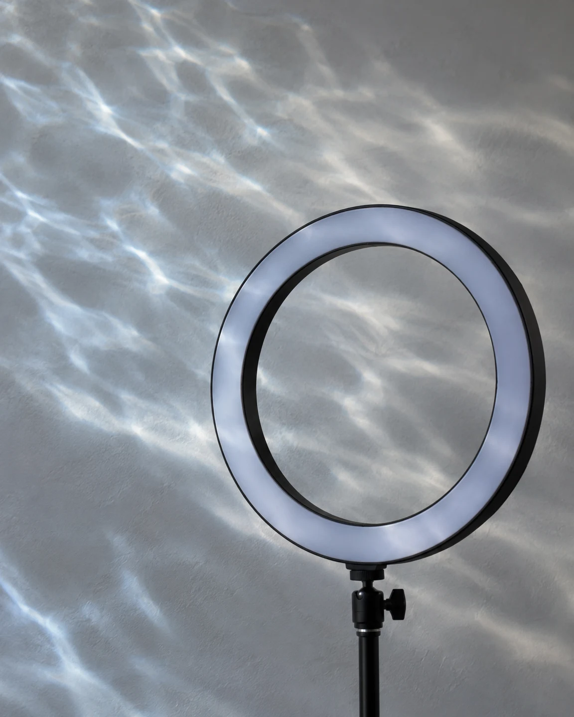 A black ring light on a slim stand is positioned against a pale grey studio wall, with blue-white water-ripple patterns crossing the wall and visible through…