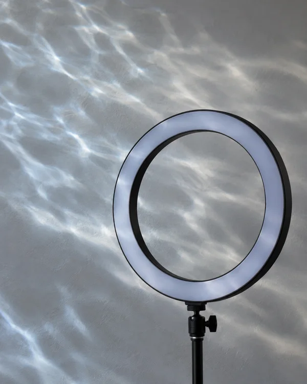 A black ring light on a slim stand is positioned against a pale grey studio wall, with blue-white water-ripple patterns crossing the wall and visible through…