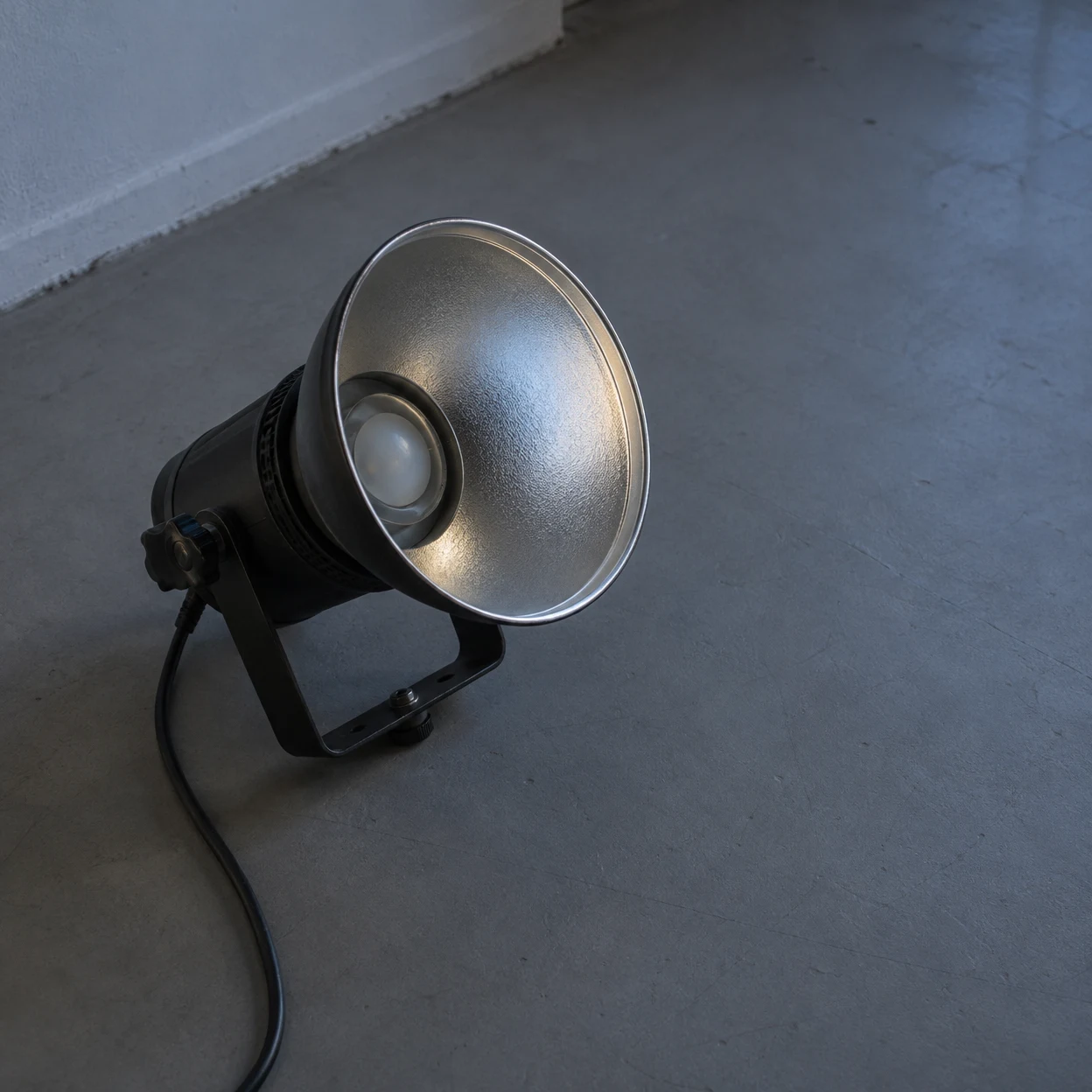 A black studio light with a broad silver reflector rests on its metal yoke on a grey floor beside a pale wall; its cable curves down toward the bottom edge…