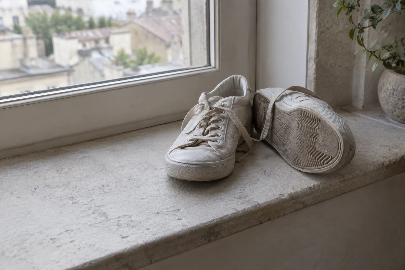 A pair of off-white canvas trainers rests on a pale stone windowsill beside a softly blurred window.