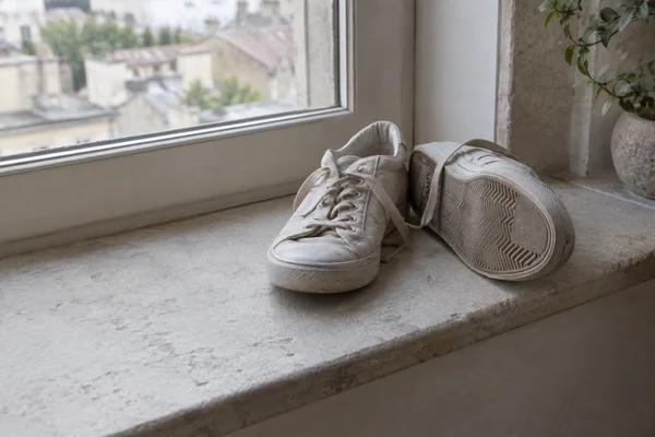 A pair of off-white canvas trainers rests on a pale stone windowsill beside a softly blurred window.