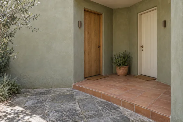 A recessed courtyard entrance with a wooden door and an off-white door on perpendicular sage-green walls, opening onto a terracotta-tiled landing above…