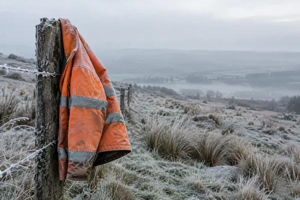 An empty orange waterproof work jacket with grey reflective bands, dusted with frost, hangs from a weathered wooden fence post in a quiet upland field.