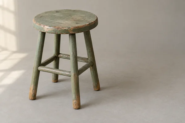 A worn round wooden stool with faded sage-green paint stands on a warm-gray floor against a matching background.