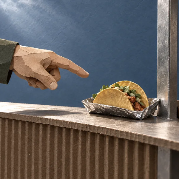 A layered-paper hand points toward two tacos in a crinkled foil tray on a corrugated counter, against a slate-blue background with a metal stall upright at…