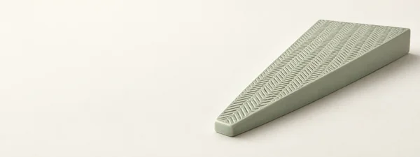 A pale celadon ceramic wedge with a densely engraved herringbone-like top rests on a warm ivory surface, leaving open space to the left.