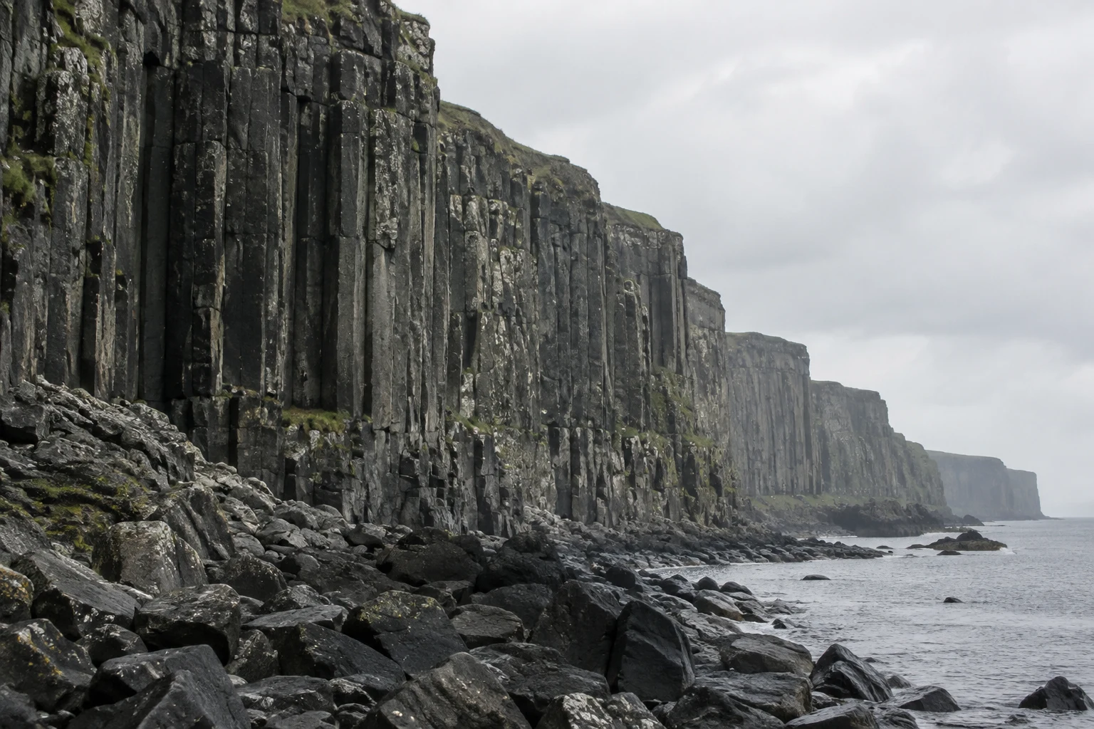 A dark basalt sea cliff with vertical columnar ribs recedes into sea haze above a rocky shoreline, with wet boulders, grey water and pale overcast sky.