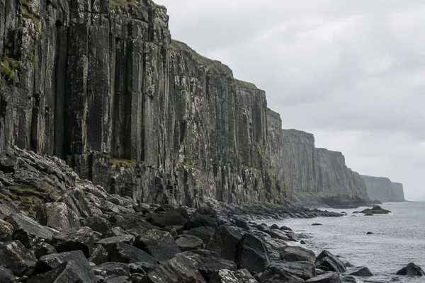 A dark basalt sea cliff with vertical columnar ribs recedes into sea haze above a rocky shoreline, with wet boulders, grey water and pale overcast sky.