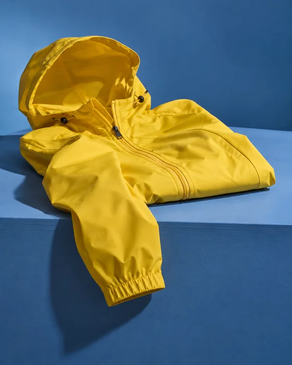 A sunflower-yellow hooded raincoat with a visible zipper lies folded on a blue tabletop, one sleeve draped over the front edge and ending in a cuff.