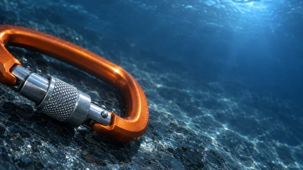 A large orange climbing carabiner with a silver knurled locking sleeve lies diagonally on a dark, textured blue floor beneath clear water, with bubbles beside…