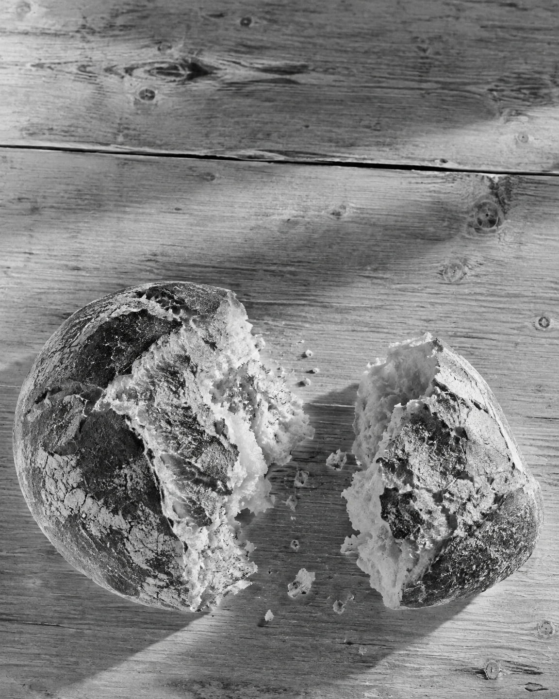 A rustic round loaf torn into two uneven pieces sits on a weathered wooden tabletop, with the torn interiors facing a narrow gap and crumbs scattered nearby.