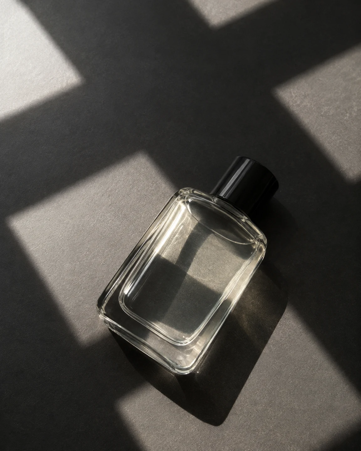 A clear rectangular glass bottle with a black cap lies diagonally on a charcoal tabletop, crossed by large patches of light and shadow.