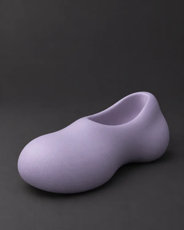 A pale lilac, rounded slip-on shoe with a recessed opening sits at an angle on a charcoal floor, with ample empty space above it.