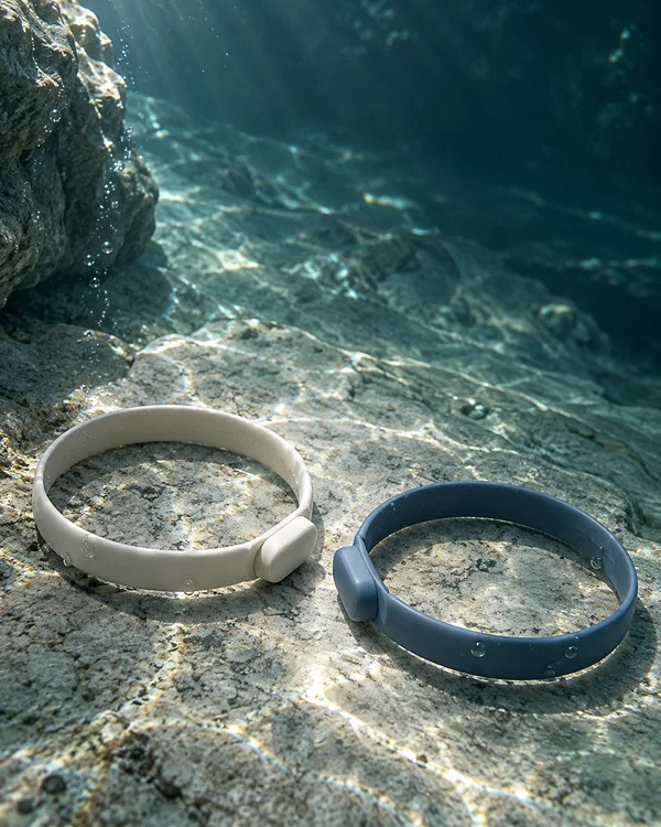 Two smooth silicone loop bracelets, one ivory and one slate blue, rest on a rocky underwater seabed with their chunky connection tabs facing each other…