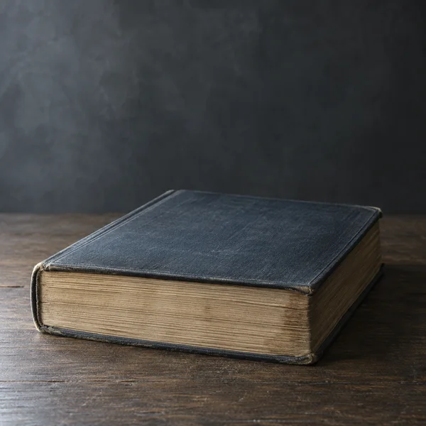 A thick, closed dark blue clothbound book with worn corners and aged cream page edges rests on a dark wooden table, lit from the left against a softly textured…