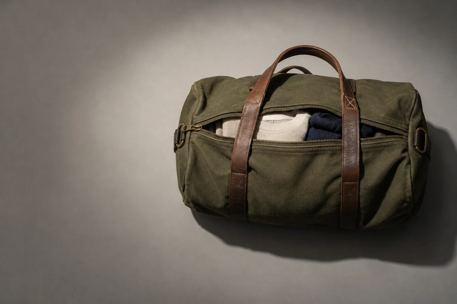 An olive canvas duffel lies horizontally on a warm-grey surface, with brown leather handles across it and a zipper opening that reveals a cream sweatshirt and…