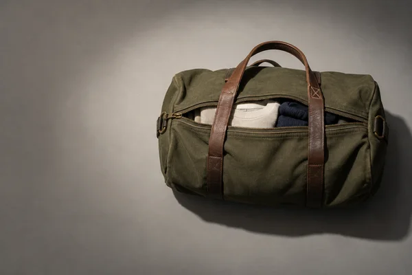 An olive canvas duffel lies horizontally on a warm-grey surface, with brown leather handles across it and a zipper opening that reveals a cream sweatshirt and…