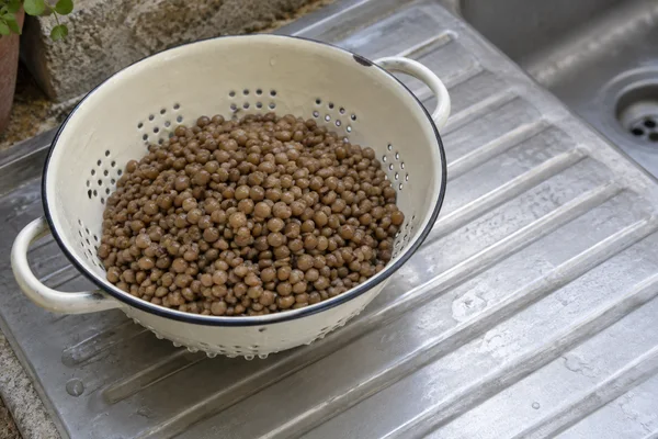 A cream enamel colander filled with brown lentils sits on a ribbed metal draining board beside a sink basin.