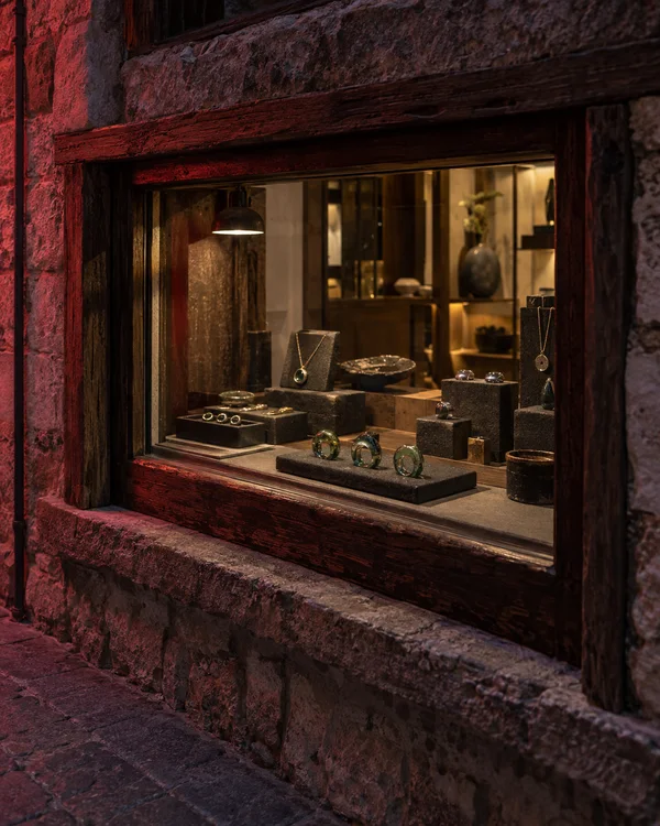 A warmly lit jewellery kiosk sits behind a weathered timber display window set into a rough stone wall, with three green glass rings arranged on a dark tray…