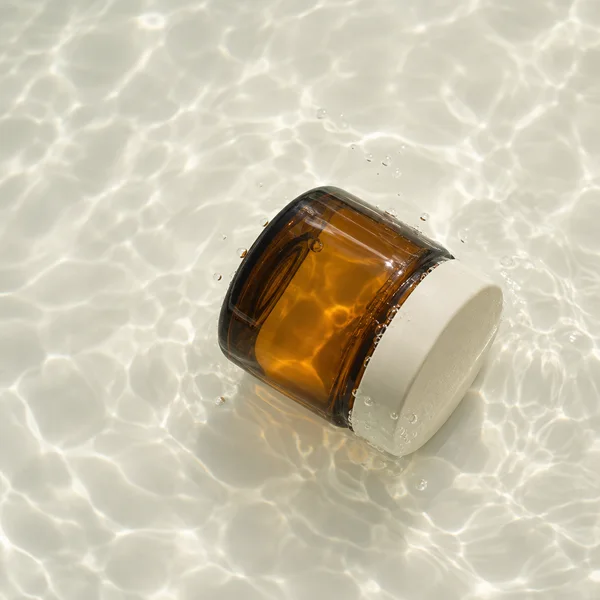 A squat amber-glass cosmetic jar with an ivory lid lies diagonally on a pale floor beneath clear water, with bright rippling light patterns and bubbles around…