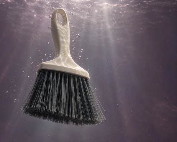 An ivory hand broom with a charcoal bristle fan hangs upright in dark, plum-grey water, surrounded by small bubbles.